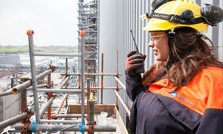 Image of a woman wearing a hard hat and using a walkie talkie while standing on scaffolding