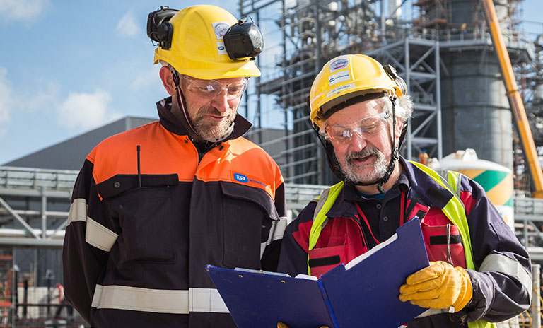 Photograph of two men with beards and wearing hard hats. They are looking at the contents inside of a blue folder. There is an energy plant in the background