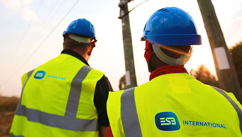 Photograph of two men with their backs to the camera and looking at power lines. They are wearing hard hats and high vis vests with the ESB International logo on the back