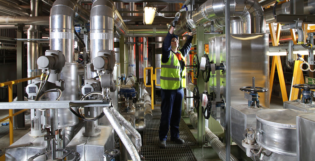 Photograph of a man wearing a high vis vest. He is reaching up to fix some pipes in a room with lots of machinery
