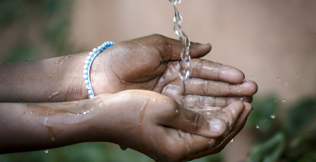 Close up photograph of brown hands cupping some pouring water. There is a blue plastic bracelet on one wrist
