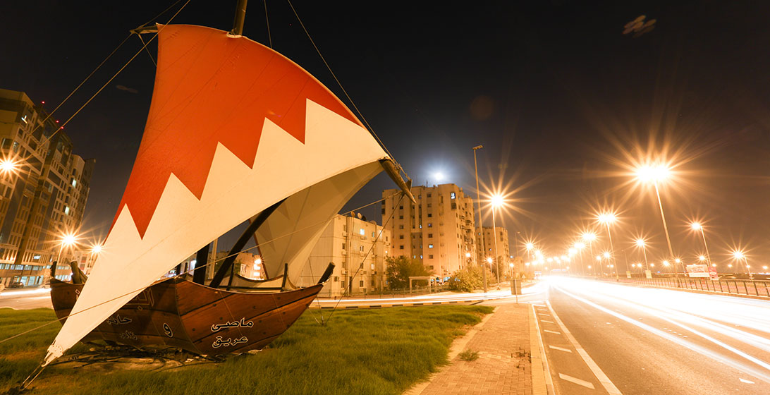 Photograph of a boat statue with a decorative red and white sail. It is on some grass next to a well lit road in a city