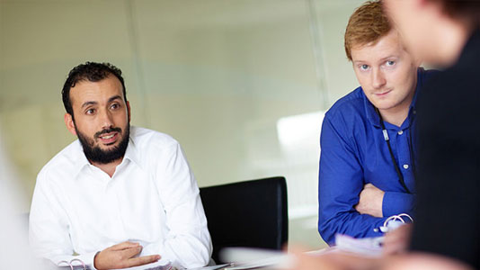 Image of two men in a meeting room listening to another man speak