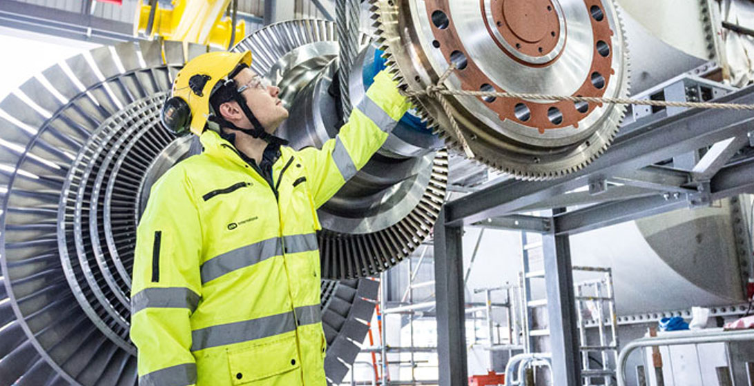 Man wearing high vis jacket and hard hat reaches up to fix a large piece of metal machinery
